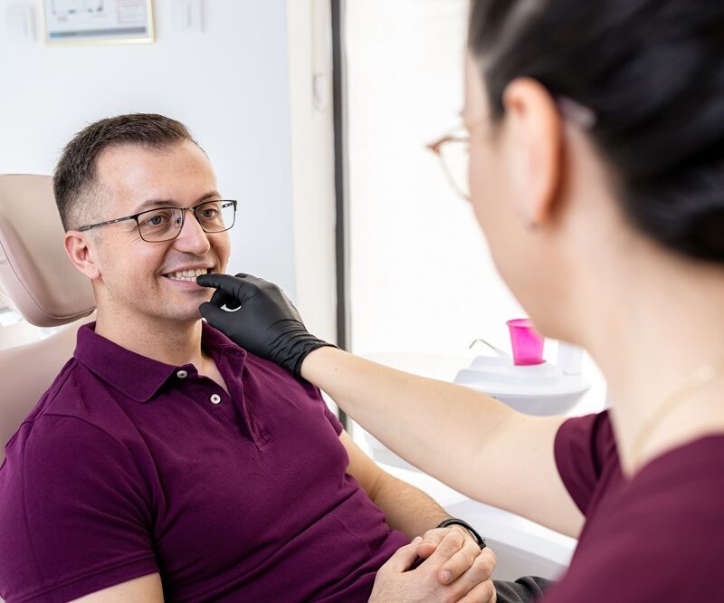 Smiling man in glasses in a dental chair as a dental team member gently checks his teeth during a veneers visit in Woodbridge, VA