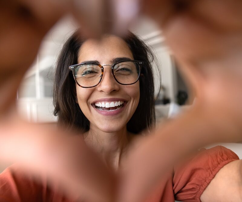 Smiling woman in glasses framing her face with her hands in a heart shape, showing the confidence of a smile makeover in Woodbridge, VA