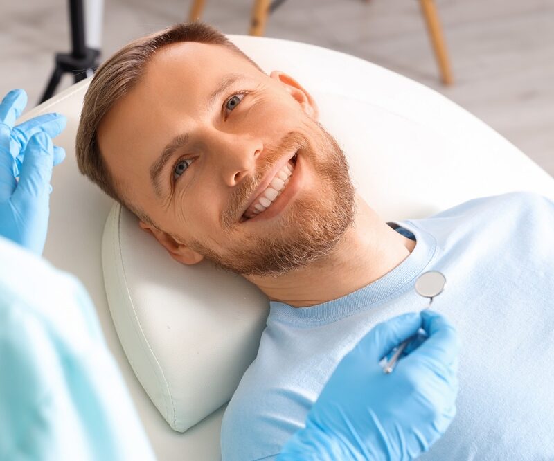 Smiling man reclined in a dental chair while a dentist in blue gloves holds a mirror during a smile makeover visit in Woodbridge, VA