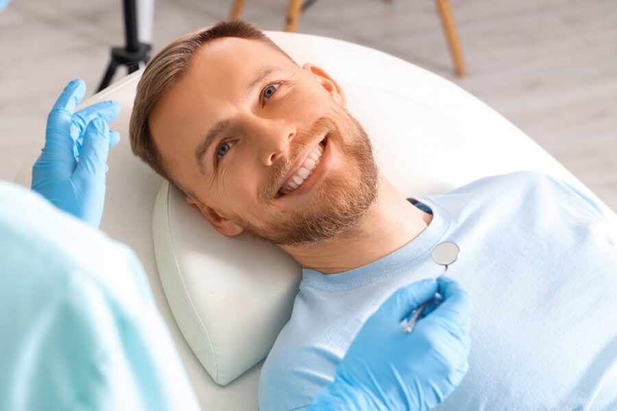 Smiling man reclined in a dental chair while a dentist in blue gloves holds a mirror during a smile makeover visit in Woodbridge, VA